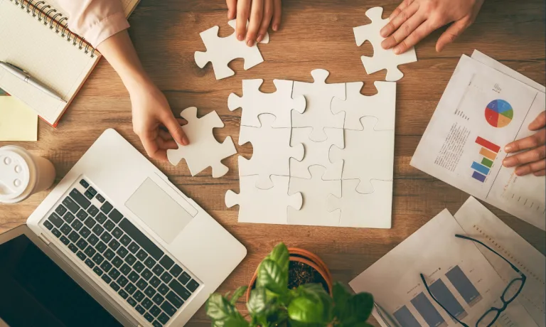 Overhead view of people collaborating to assemble puzzle pieces on a table, surrounded by a laptop, charts, and documents