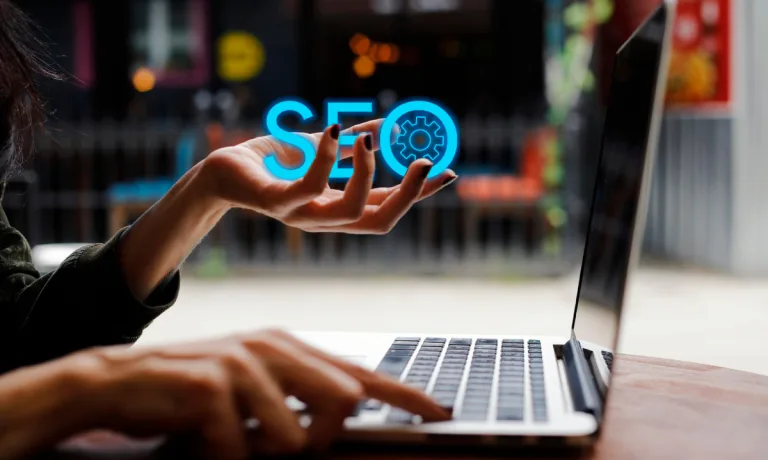 Person holding a digitally rendered 'SEO' graphic in their hand with a laptop open in front of them